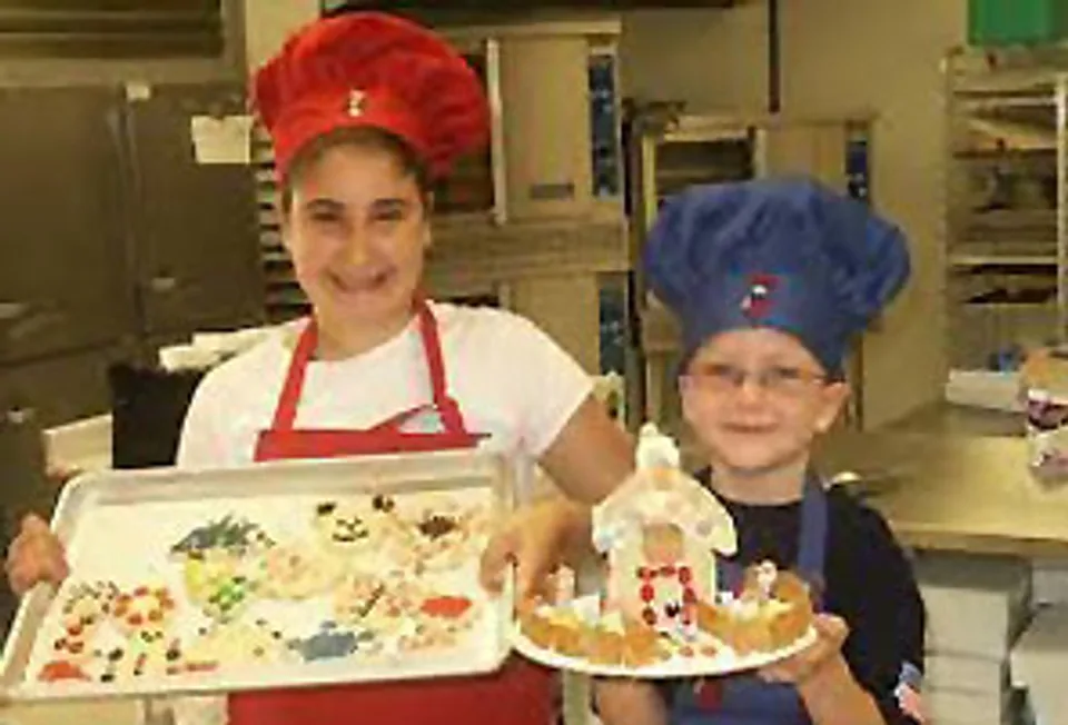Two children in chef hats proudly holding a decorated gingerbread house at an Easy Sweets baking class